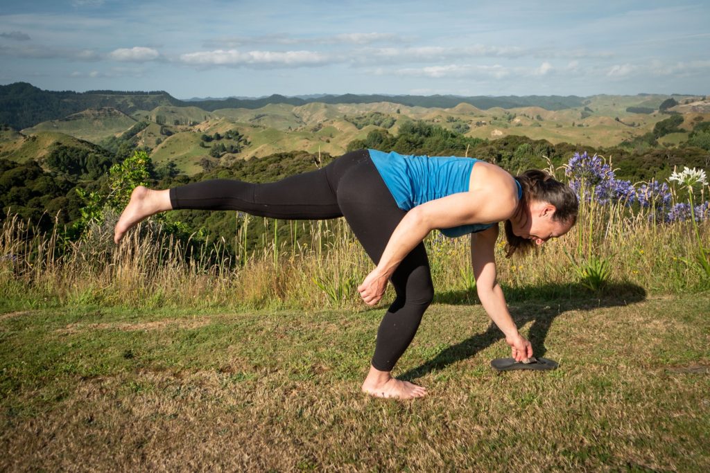 Megan reaching down to the groun with one leg lifted behind to help balance while picking up a flip flop from the ground